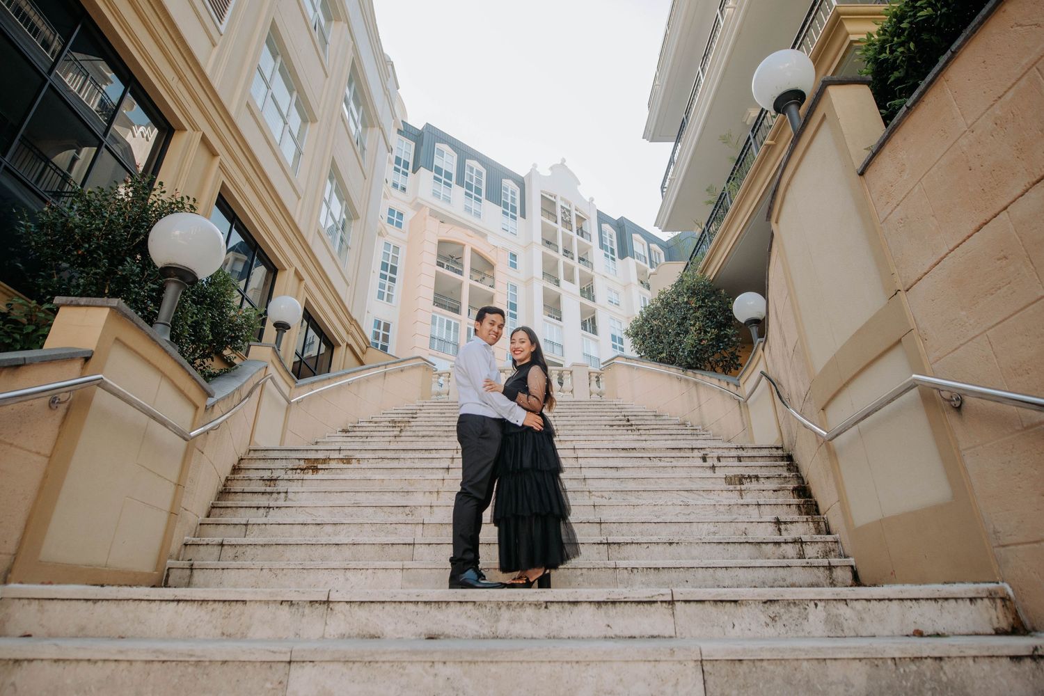 A couple poses together on elegant beige stone steps between buildings.