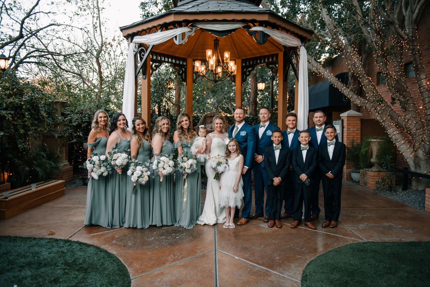 Wedding party members lined up outdoors under string lights and trees at a rustic venue.