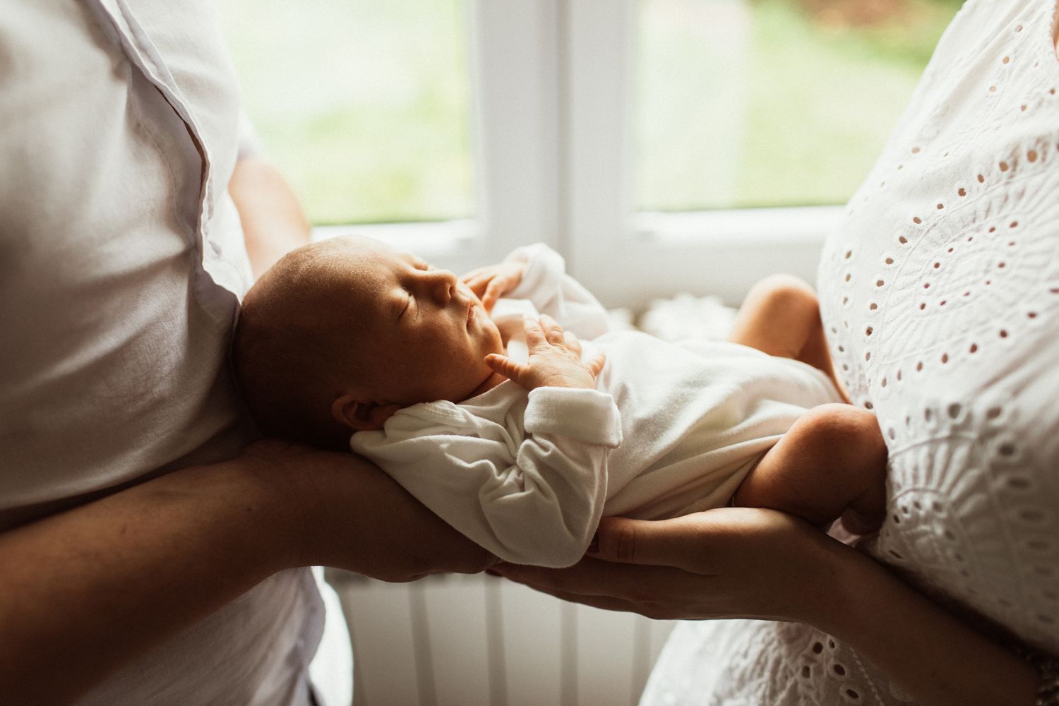 New parents cradling their sleeping newborn baby near a bright window in soft natural light.