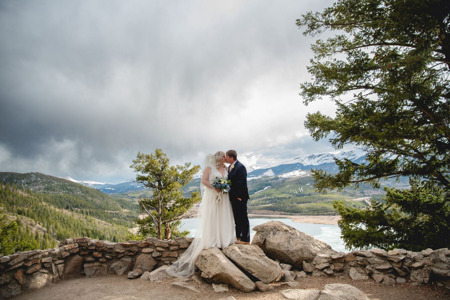 Wedding couple standing on mountain overlook with dramatic cloudy sky and mountain vista.