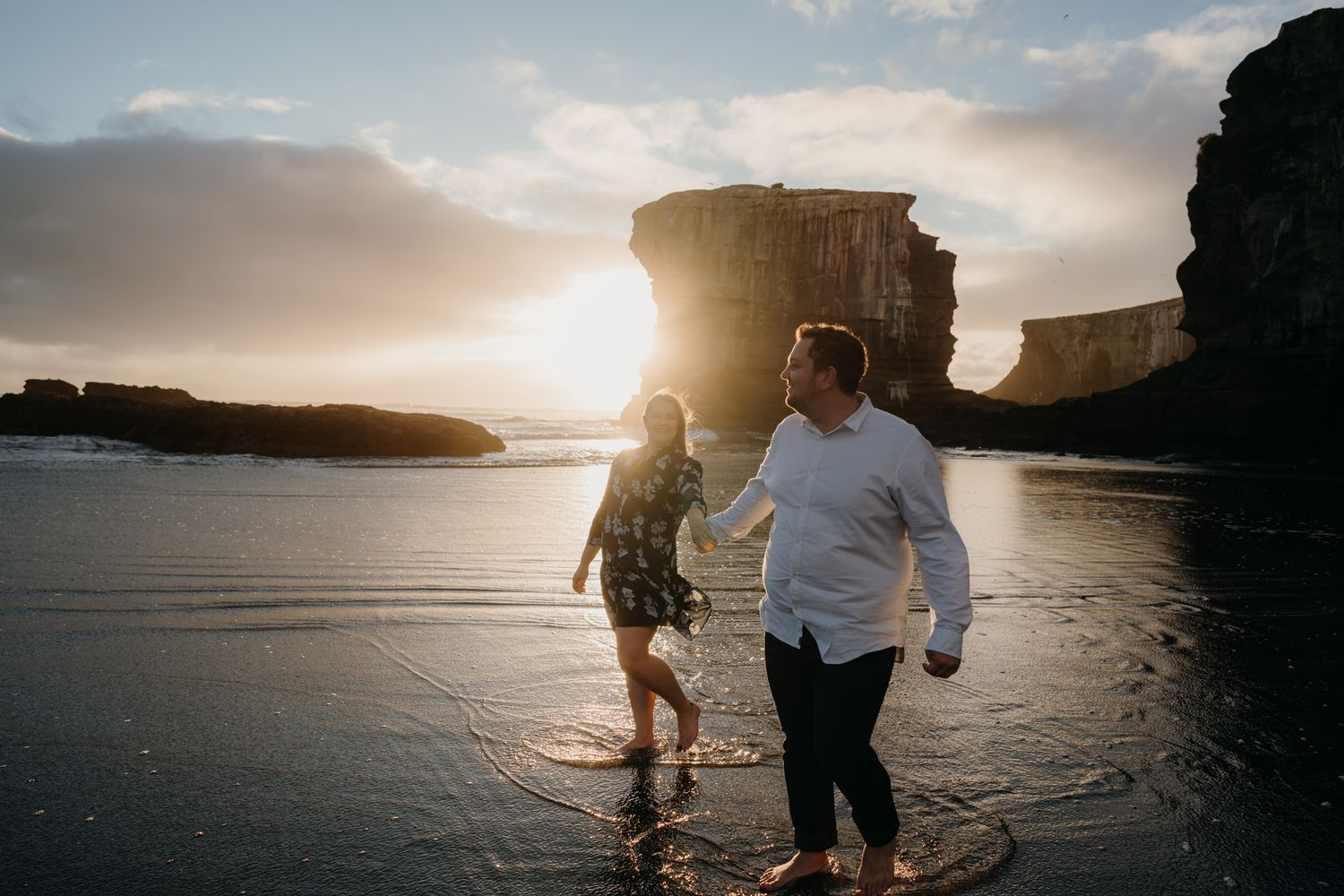 engaged couple walking along the beach holding hands at sunset with a large rock face and sunset in the background