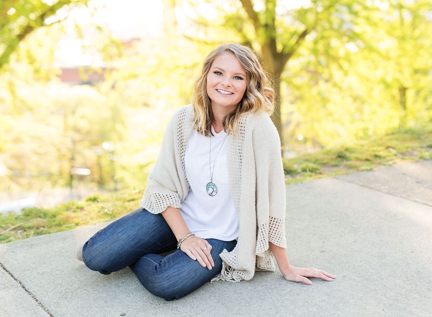 A person in a cream cardigan sits casually on a path with autumn foliage in the background.