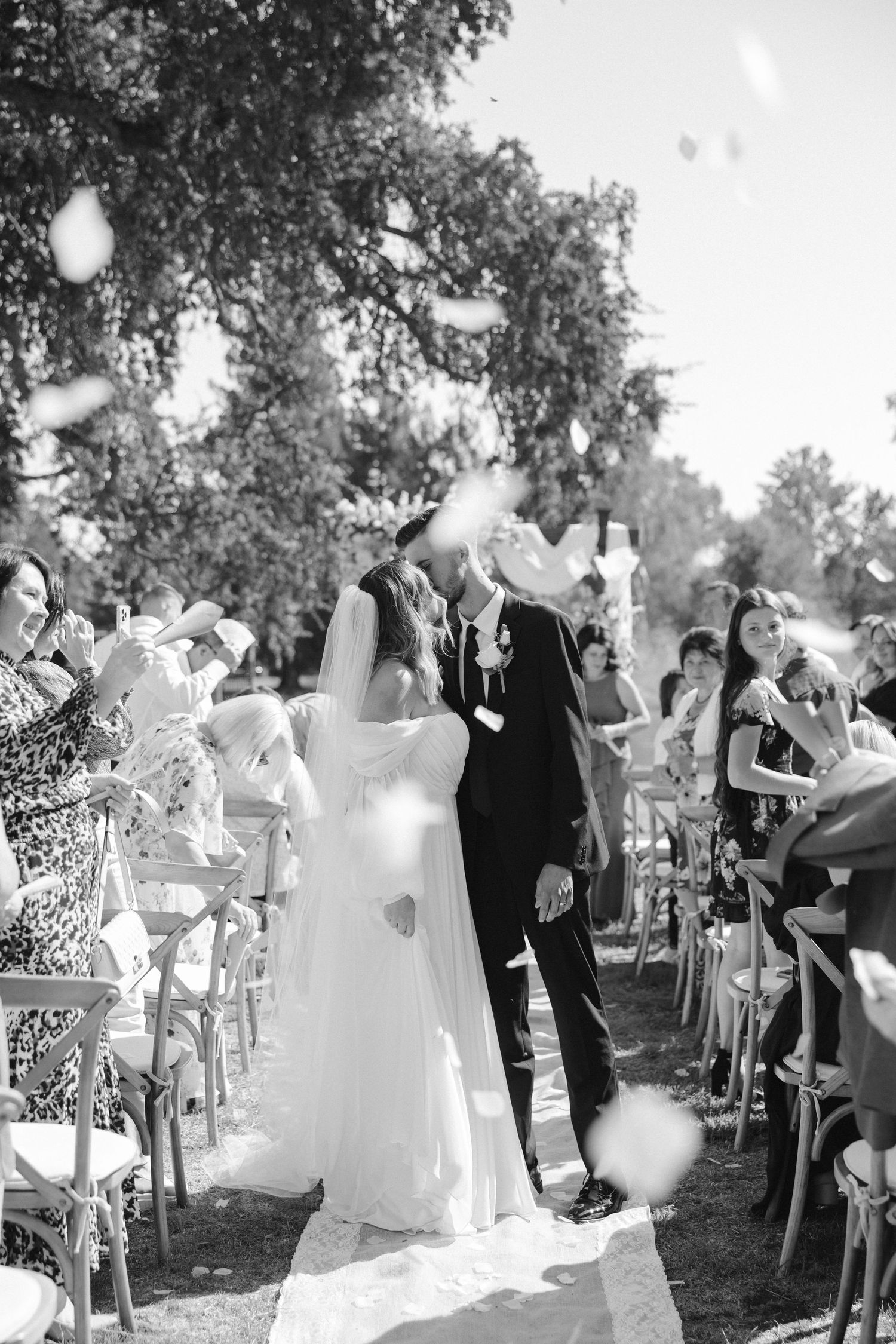 A black and white wedding ceremony photo with floating bubbles and guests seated under trees.