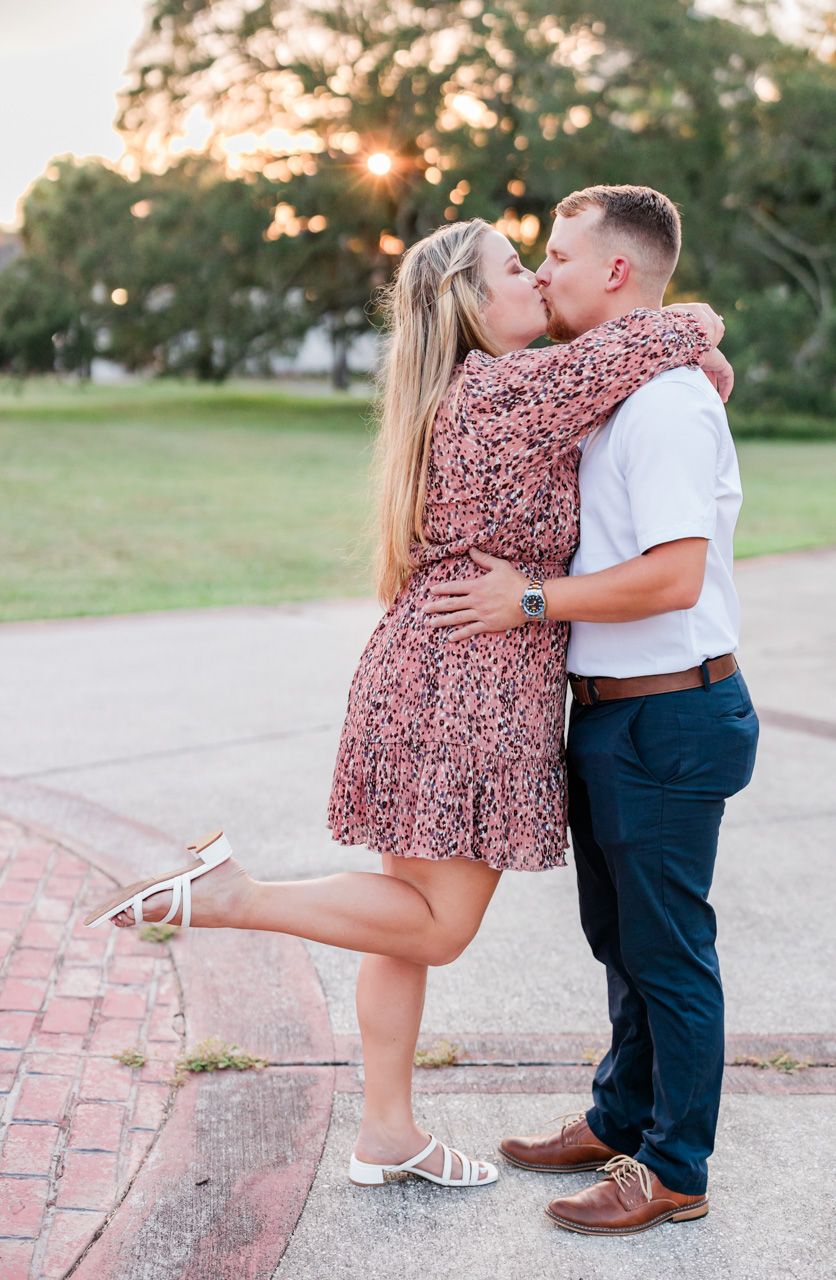 Romantic sunset kiss with woman in floral dress lifting leg during engagement photoshoot.