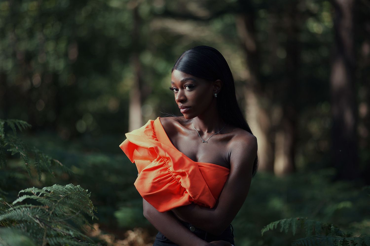 Portrait of black model in surrounded by foilage in the woods. She is wearing an off the shoulder orange top.