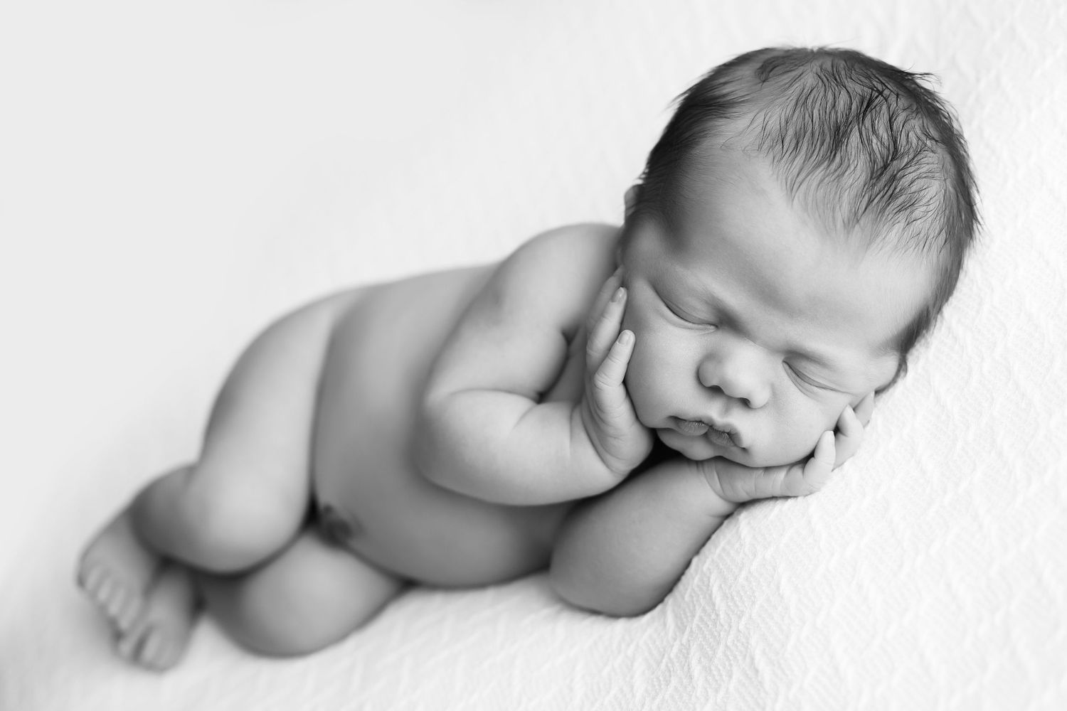 Newborn baby boy laying on a knitted white blanket.  Baby is laying on his side with his hands holding his cheeks.