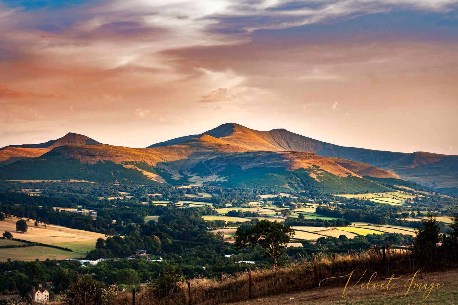Panoramic mountain landscape with rolling hills and dramatic cloudy sky at sunset.