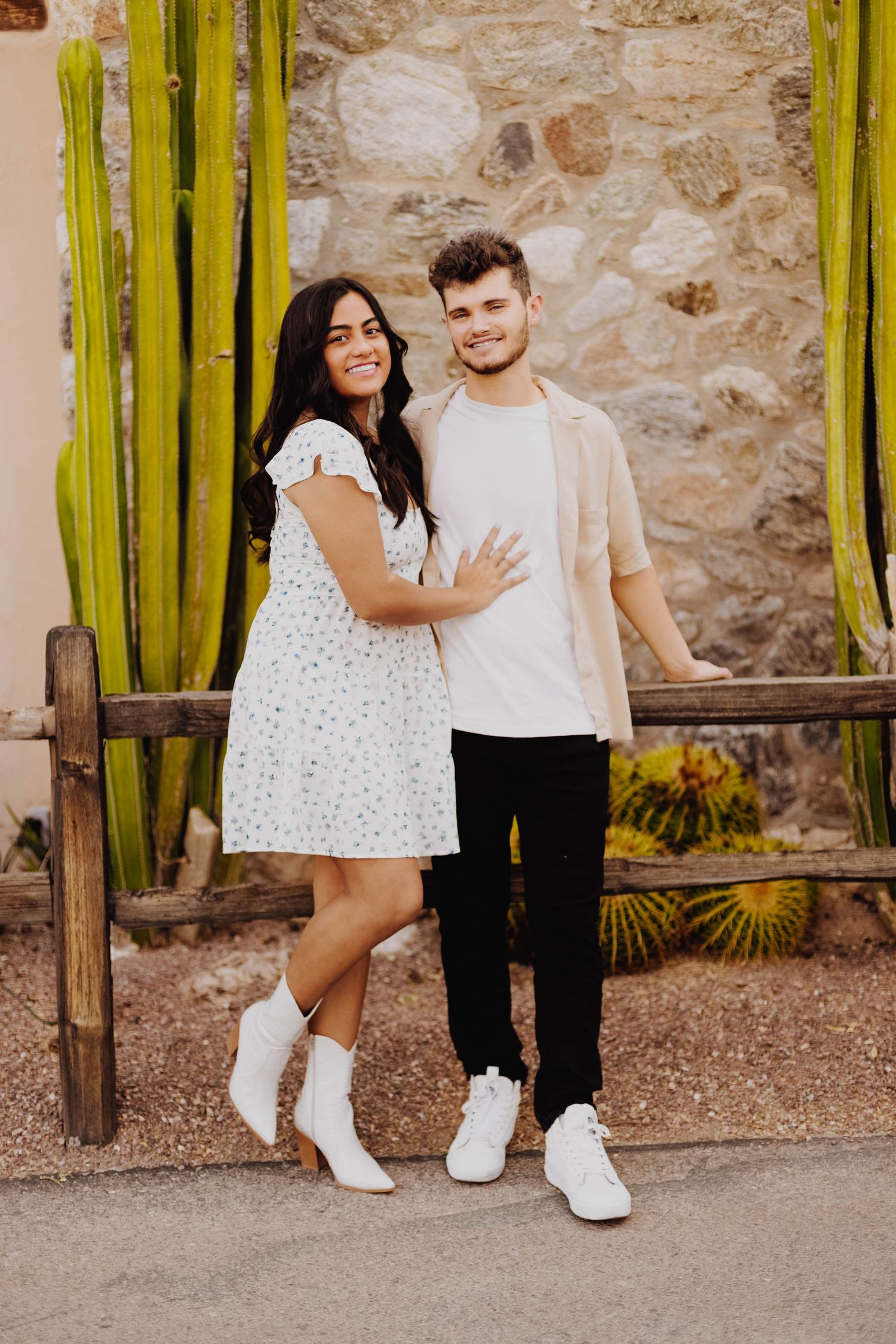 Casual couple posing together near rustic stone wall with tall cacti in background.