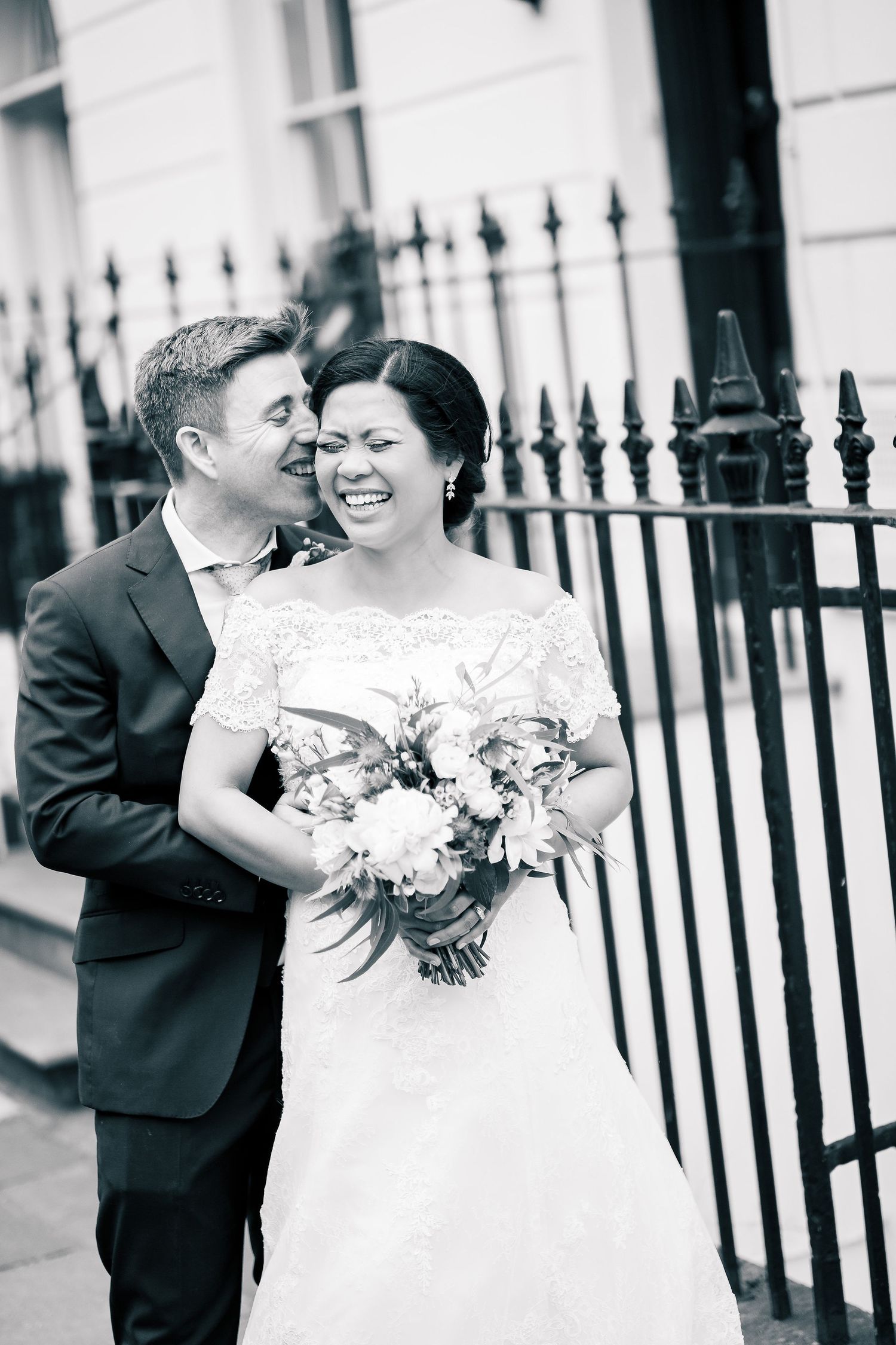 Black and white wedding photo showing a joyful moment between newlyweds next to an iron fence.