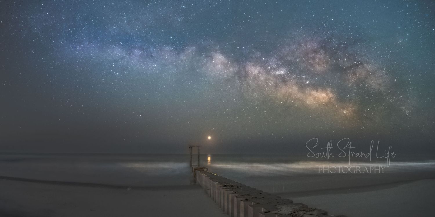 Milky Way galaxy stretches across night sky above illuminated pier extending into ocean.