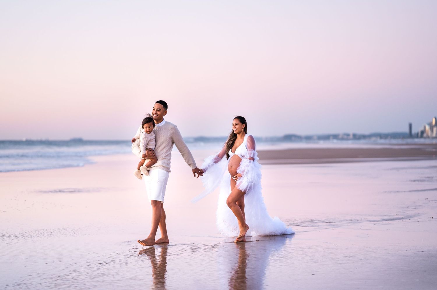 Family walking hand in hand on beach at sunset in white clothing with flowing dress.