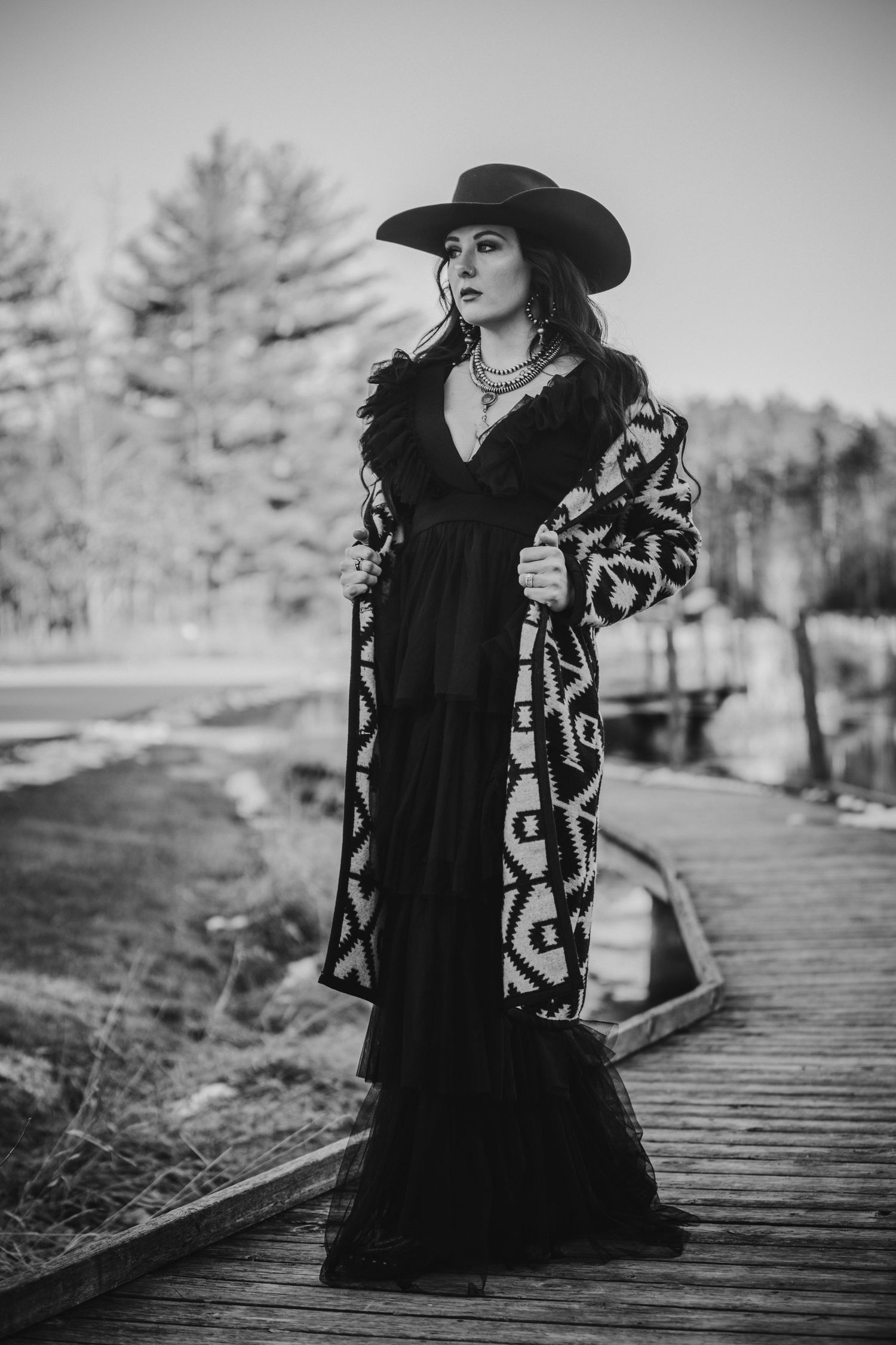 Dramatic black and white portrait of someone in a western-inspired outfit with cowboy hat and patterned coat on wooden walkway.