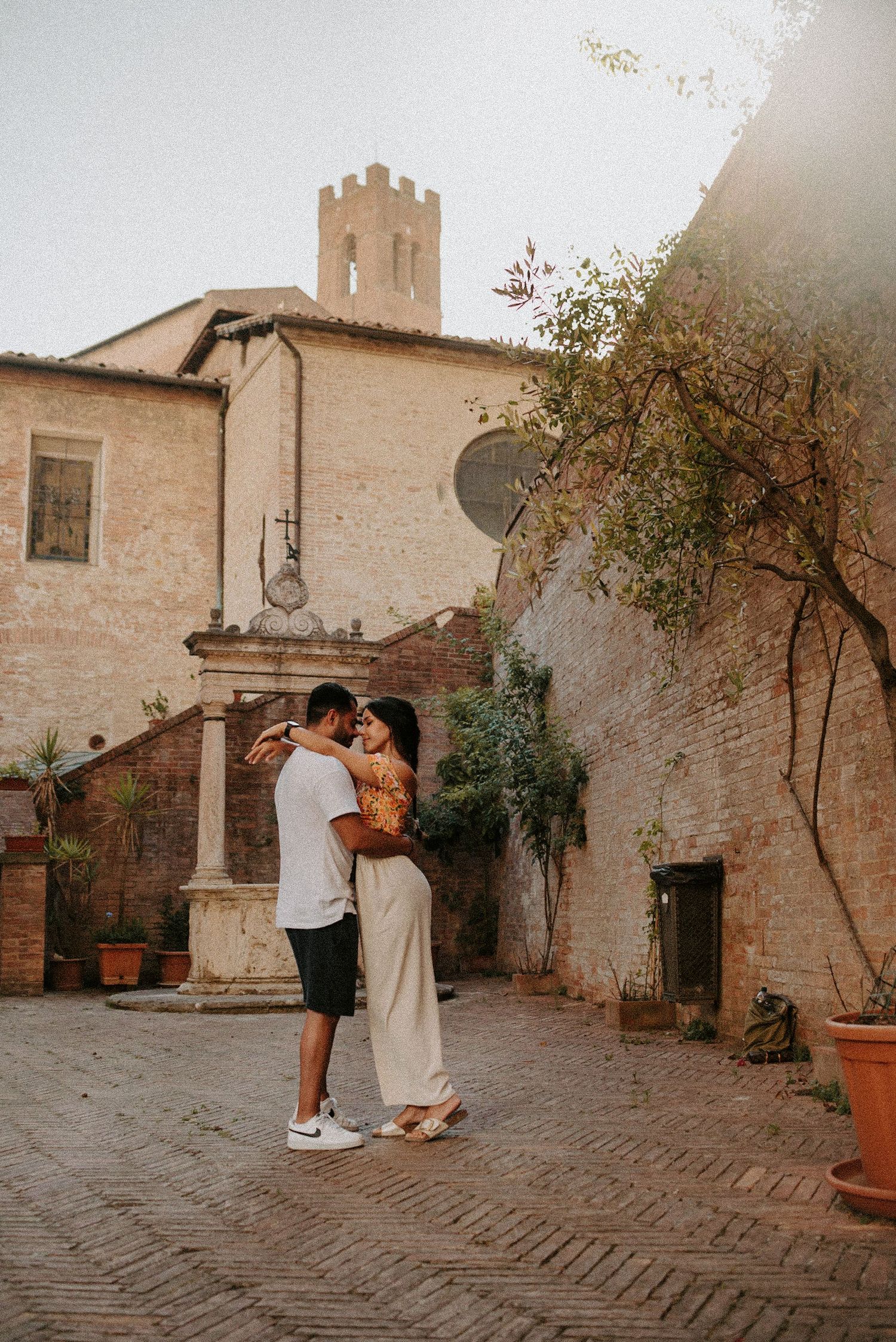 Couple embraces in courtyard of historic Italian building with tower and brick pavement.