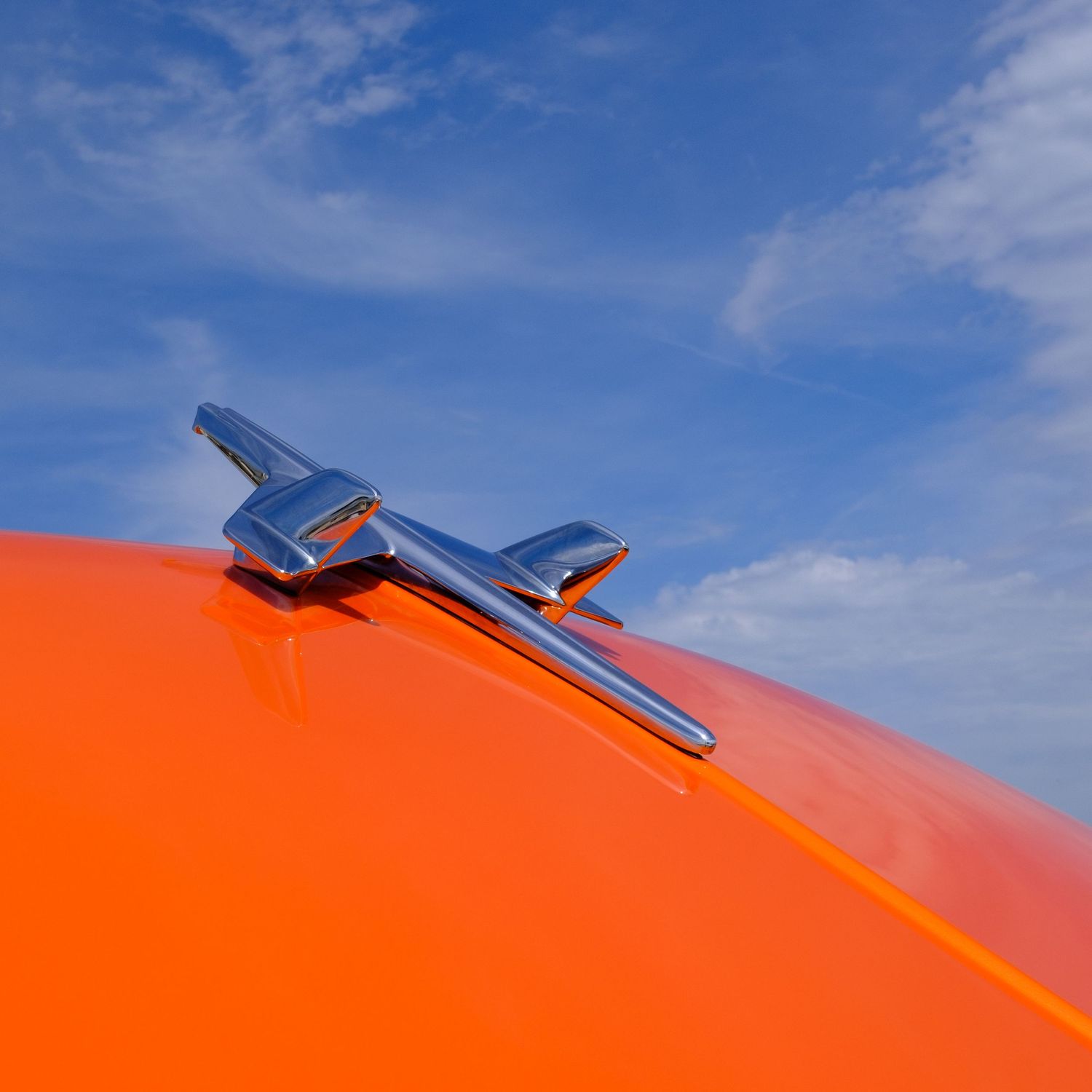 Shiny chrome hood ornament on a bright orange classic vintage car against a blue sky.