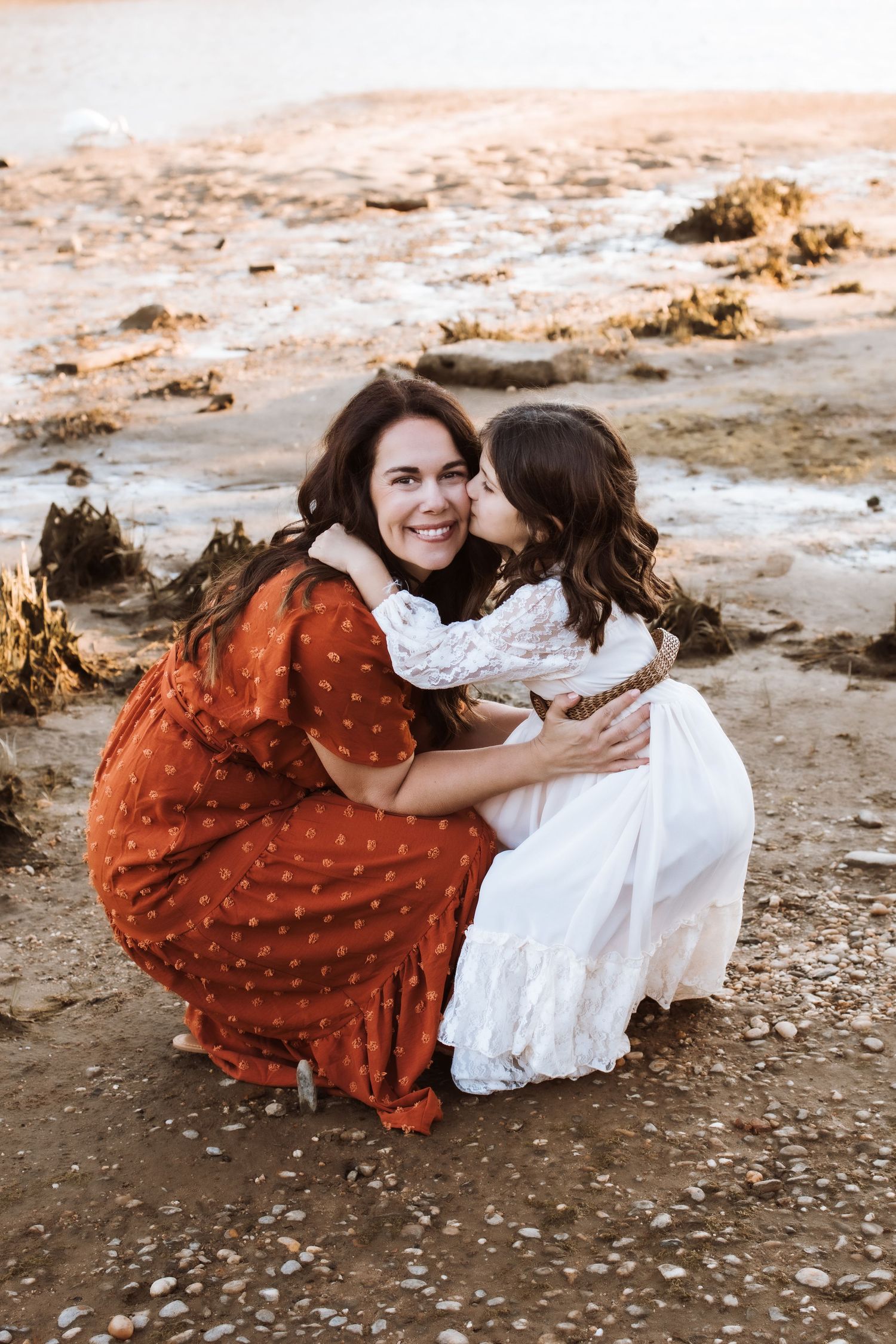 Mother crouching as daughter kisses her cheek on rocky beach at sunset.