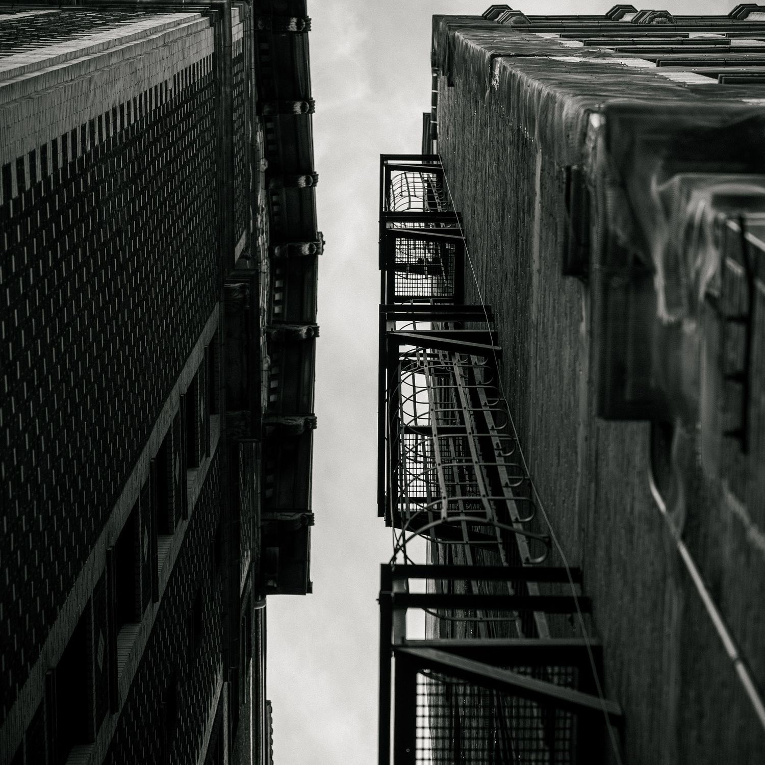 Looking up at fire escape stairs between tall buildings in black and white.