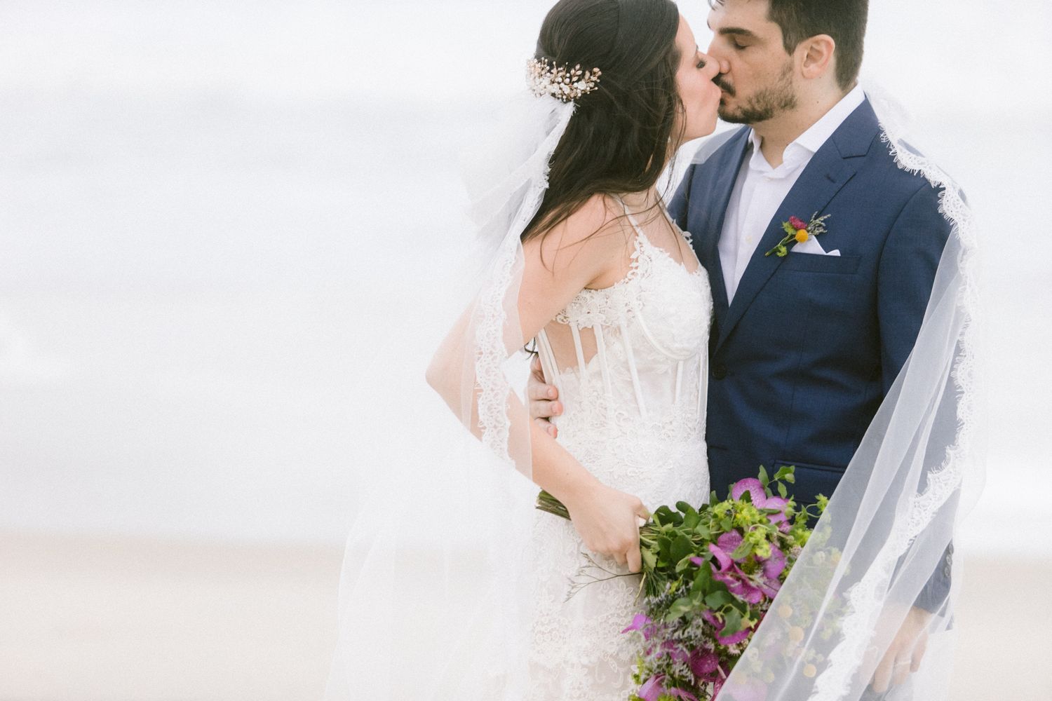 Close-up view of bridal couple in romantic beach setting with flowing veil and bouquet.