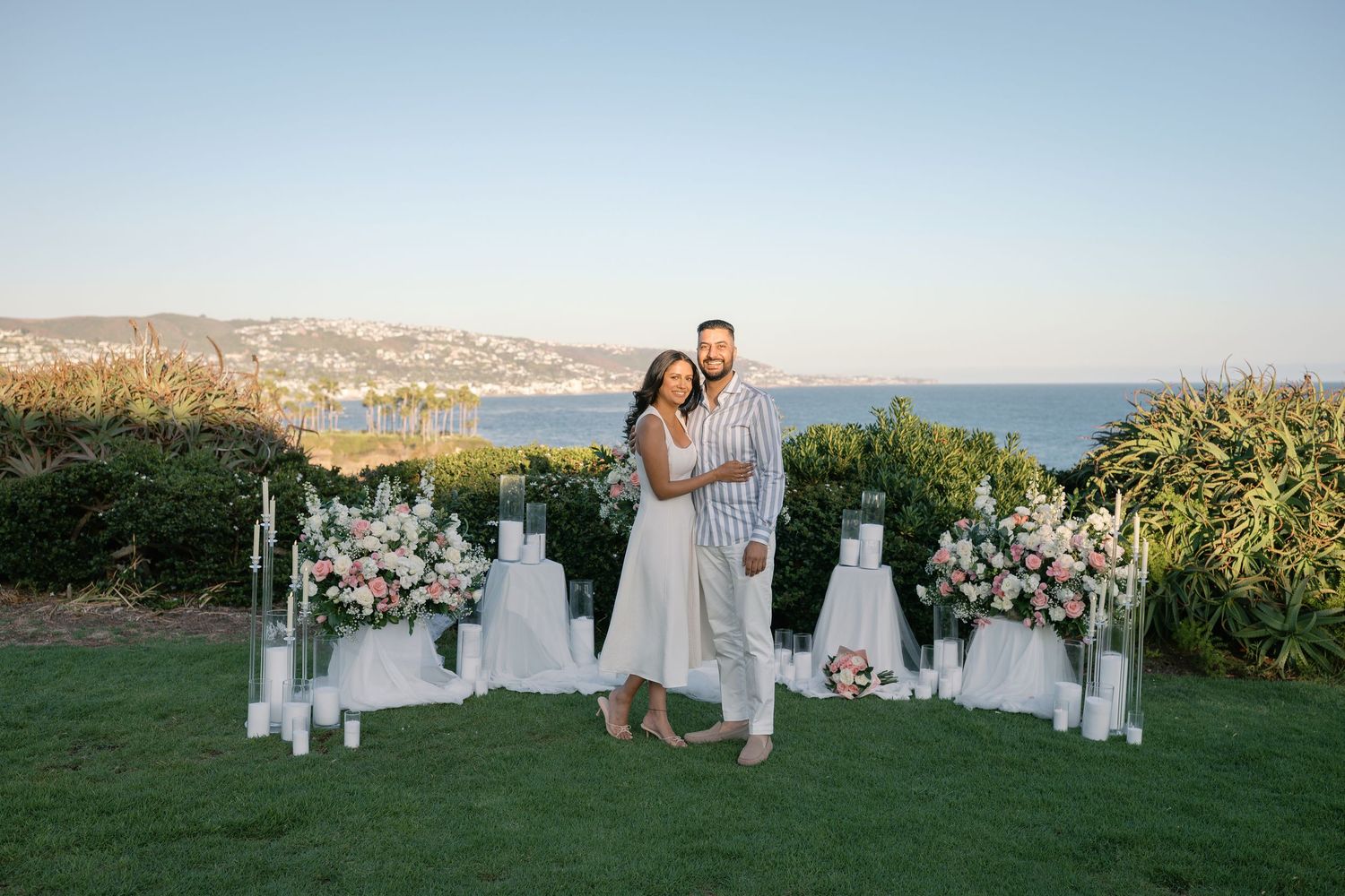Couple smiling together at outdoor engagement setup with white candles, pink flowers, and ocean view at sunset.