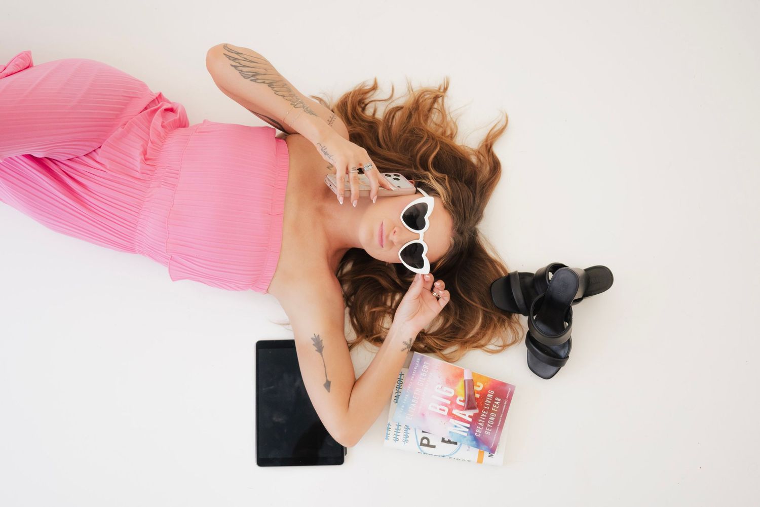 Woman in pink dress lying on white floor with sunglasses, camera, tablet, and colorful magazine.