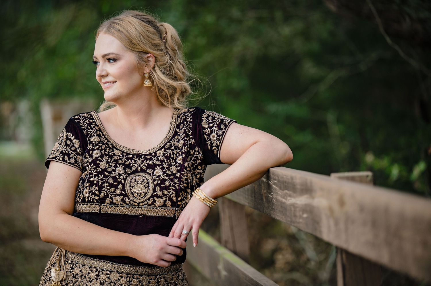 Person in embellished black and gold dress poses casually against wooden railing outdoors.