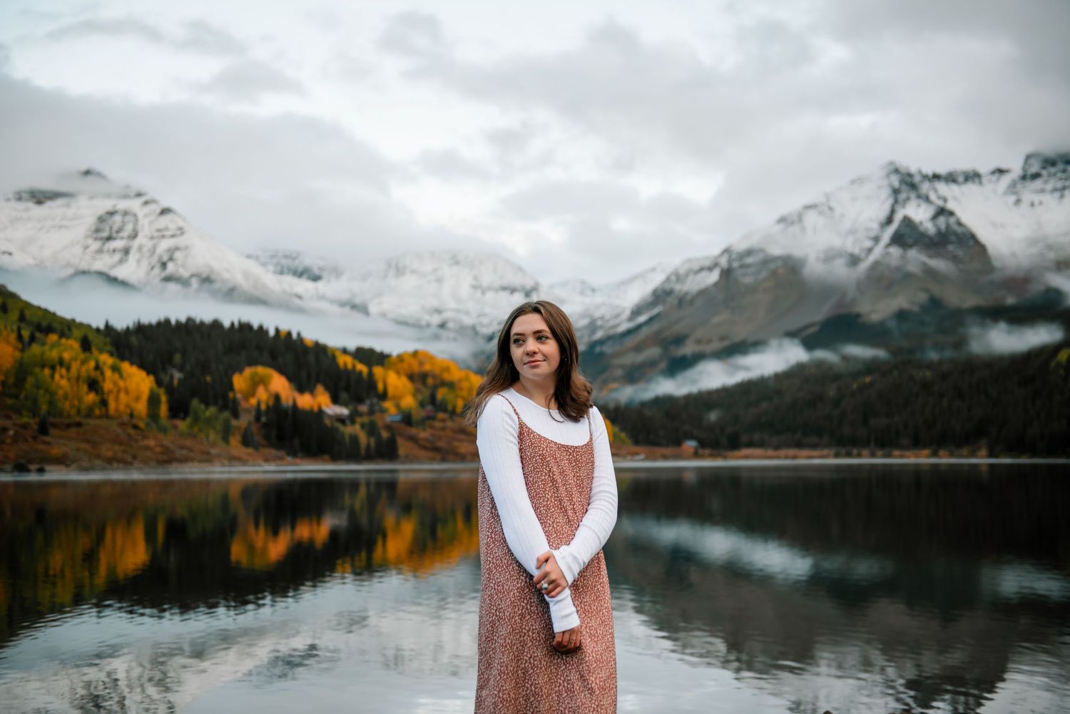 Scenic mountain lake surrounded by autumn trees and snow-capped peaks on a cloudy day.