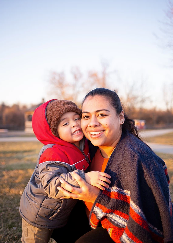 A tender outdoor moment at sunset with a small child in a red hooded jacket and someone in a patterned sweater.
