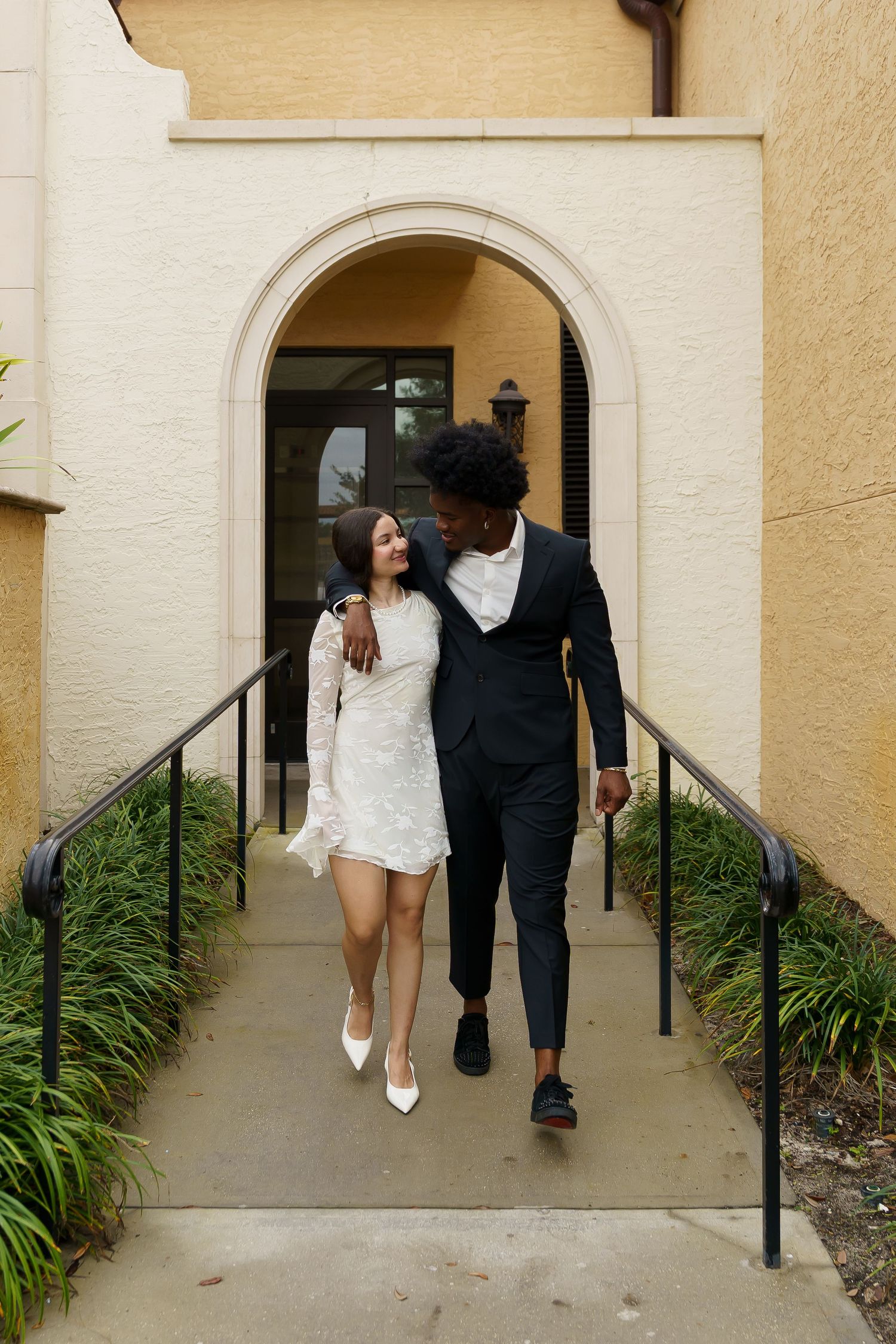 A couple in formal attire stands near a window in an elegant venue with arched ceilings and chandeliers.