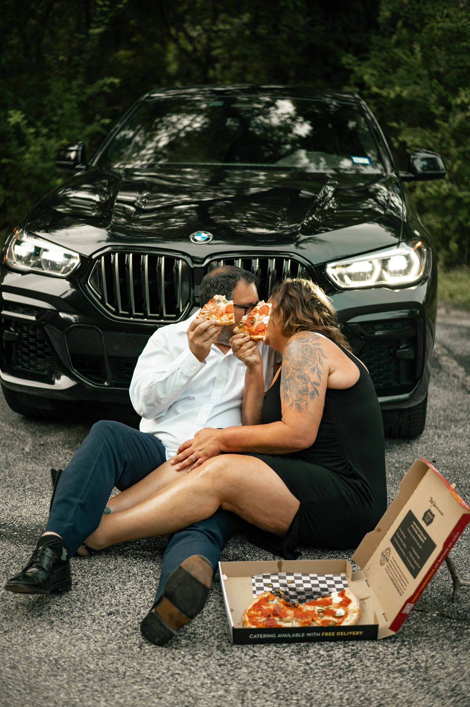 Couple sitting on ground sharing pizza in front of a black BMW SUV, with an open pizza box, surrounded by trees.