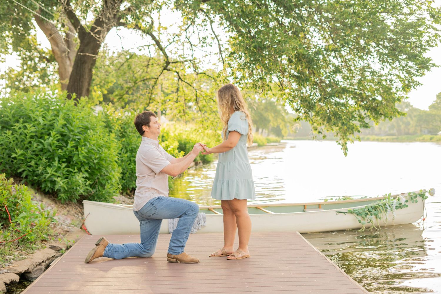Man proposes on one knee to woman on lakeside dock surrounded by lush green trees.