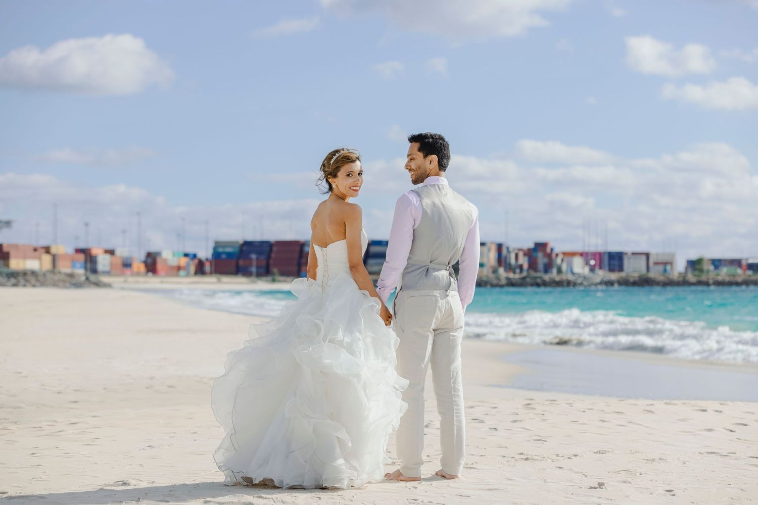 Bride and groom holding hands on a white sand beach with turquoise ocean waves and colorful buildings in the background.