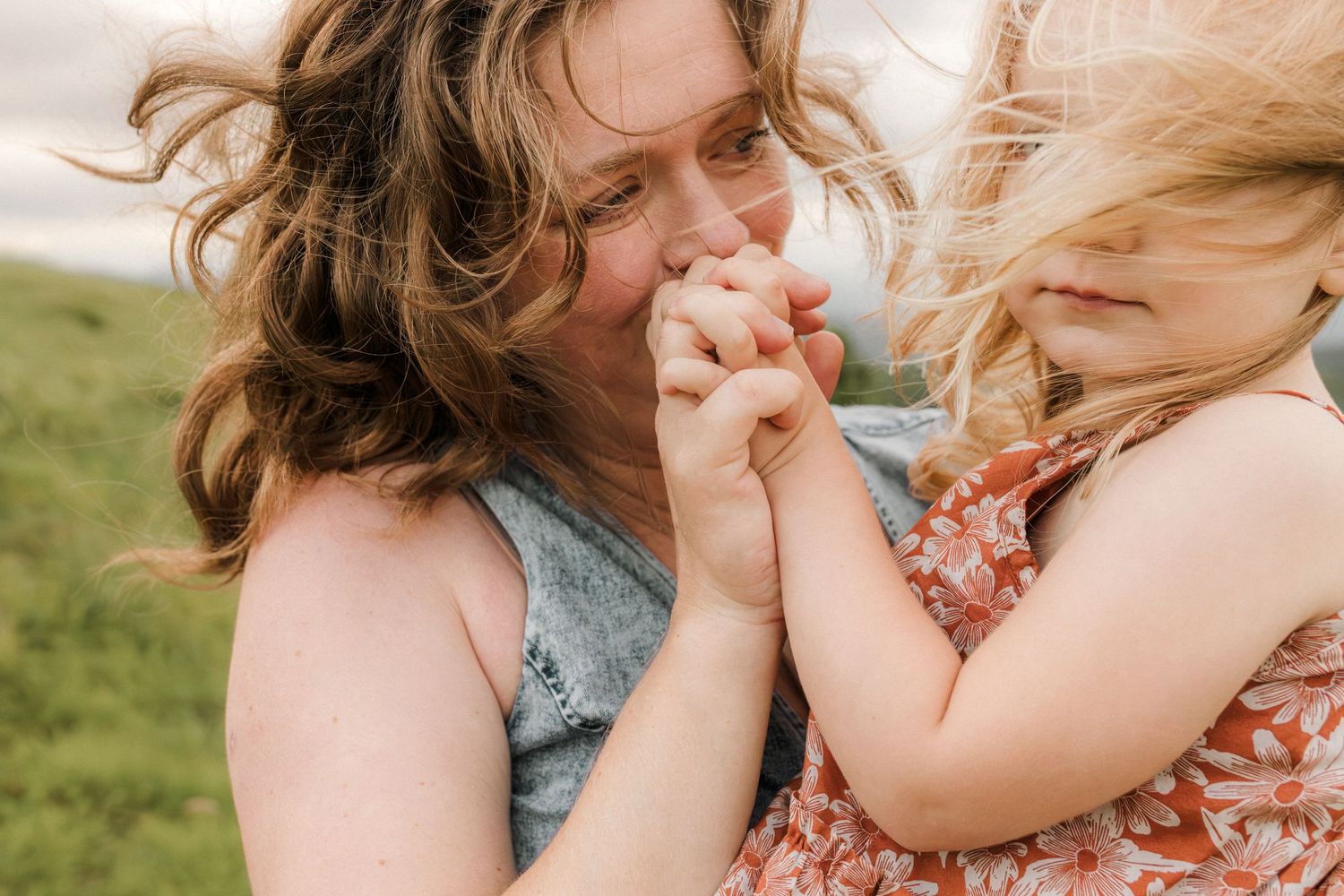 A mother tenderly kisses her young blonde daughter's hands outdoors in a heartwarming moment.