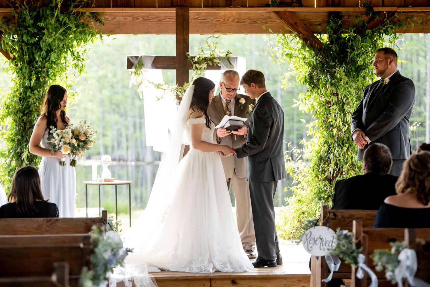 Rustic outdoor wedding ceremony under wooden pavilion decorated with greenery and natural light.