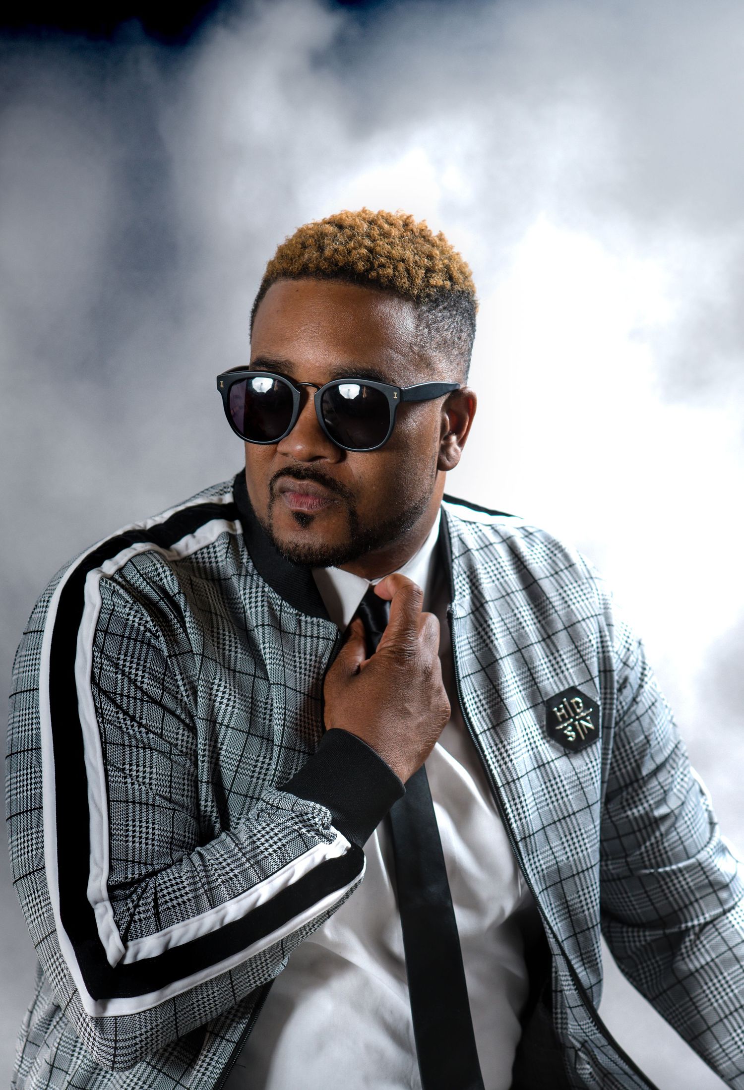 Stylish studio portrait of person in plaid jacket and sunglasses against smoky background.