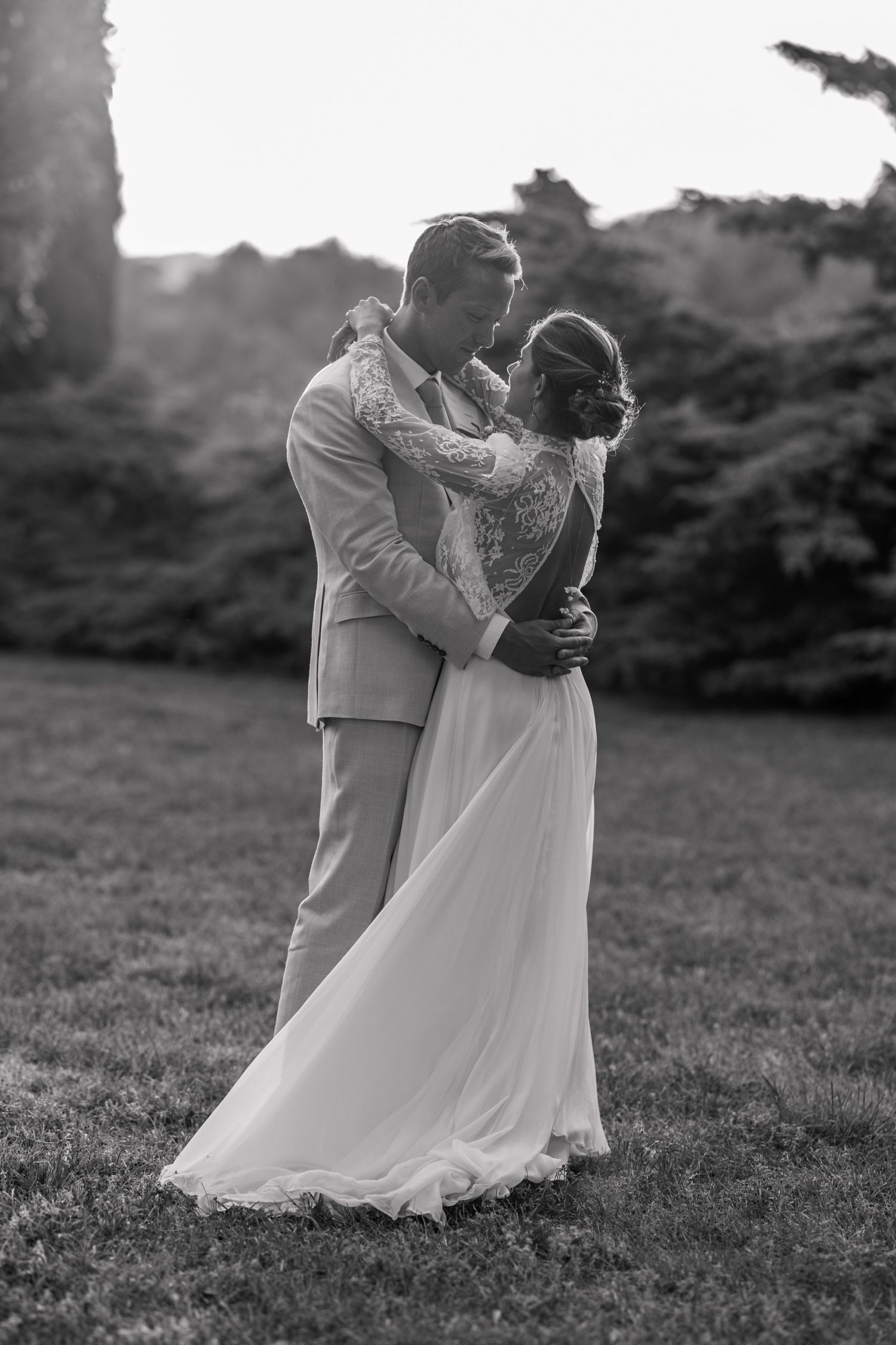 A romantic black and white wedding photo of a couple embracing on a grassy hillside in elegant attire.