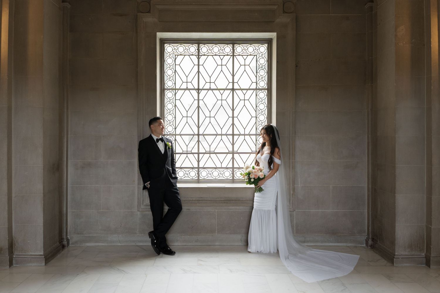 Bride and groom share a romantic moment by an ornate window in an elegant wedding venue.