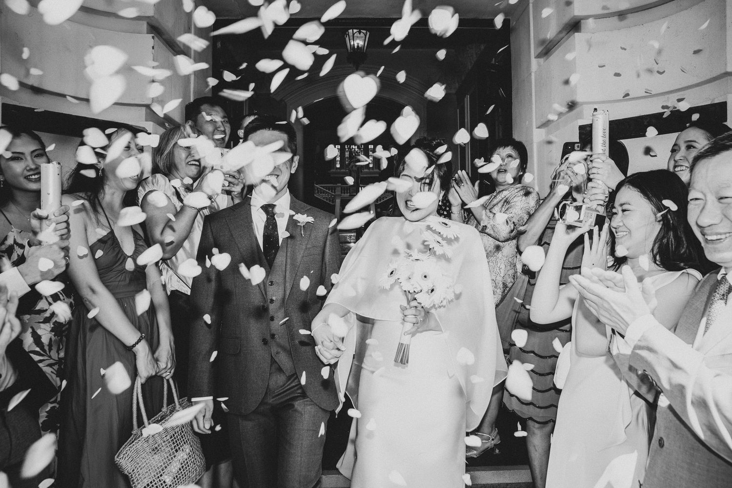 Black and white wedding photo of couple walking through confetti shower, guests celebrating with phones and flowers.