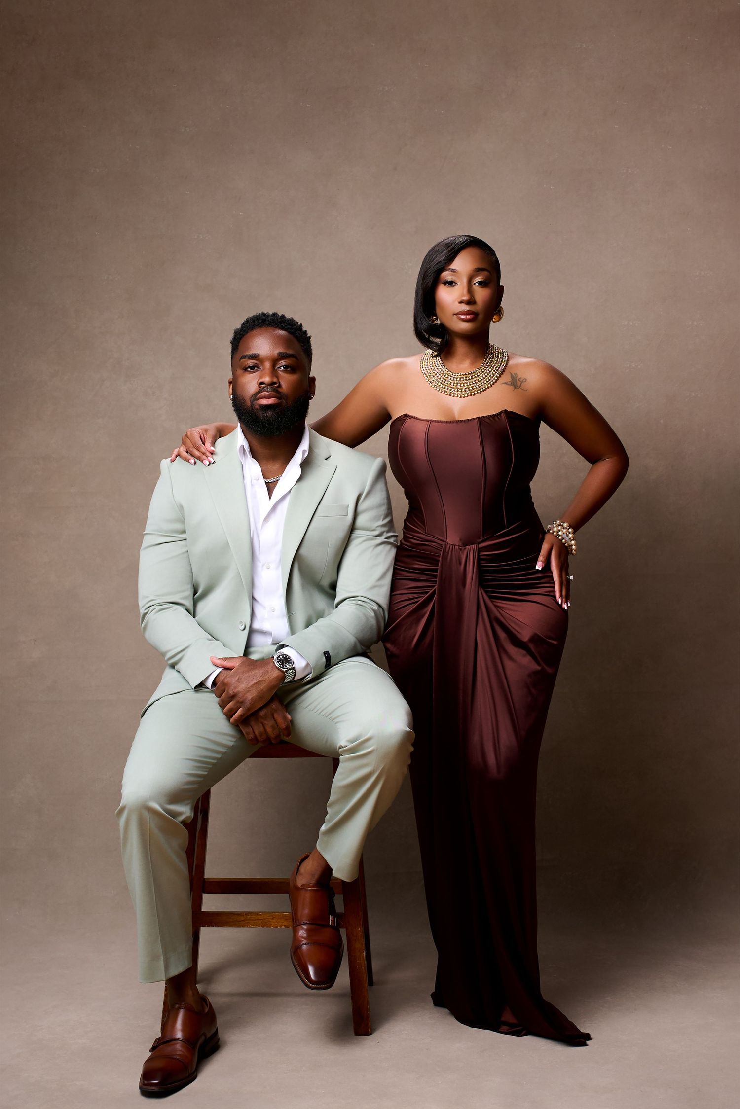 Elegant couple posing in studio, man in sage suit seated, woman in burgundy gown standing, wearing gold jewelry.
