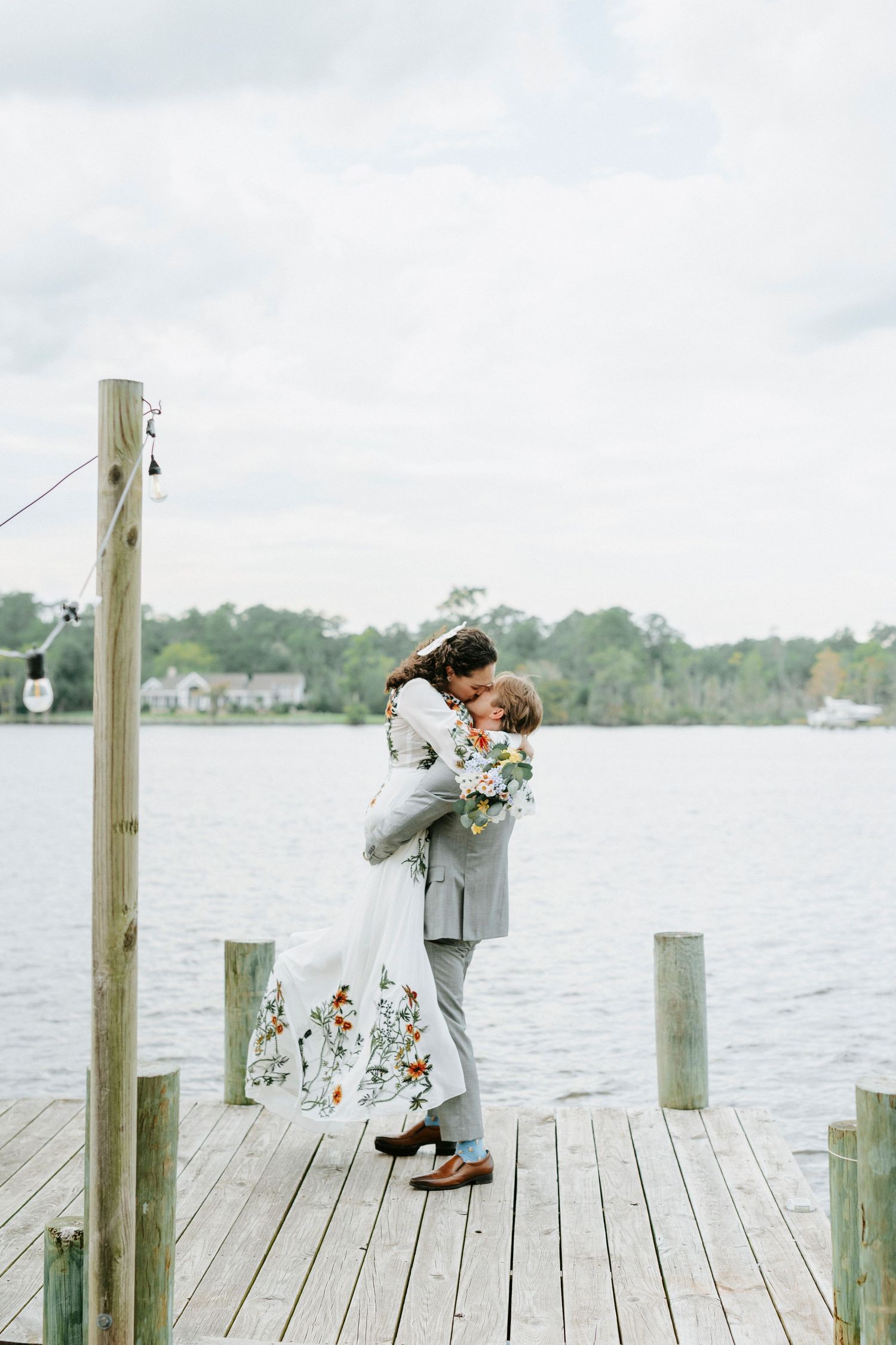 A bride and groom share a romantic kiss on a wooden dock overlooking a serene lake on their wedding day.