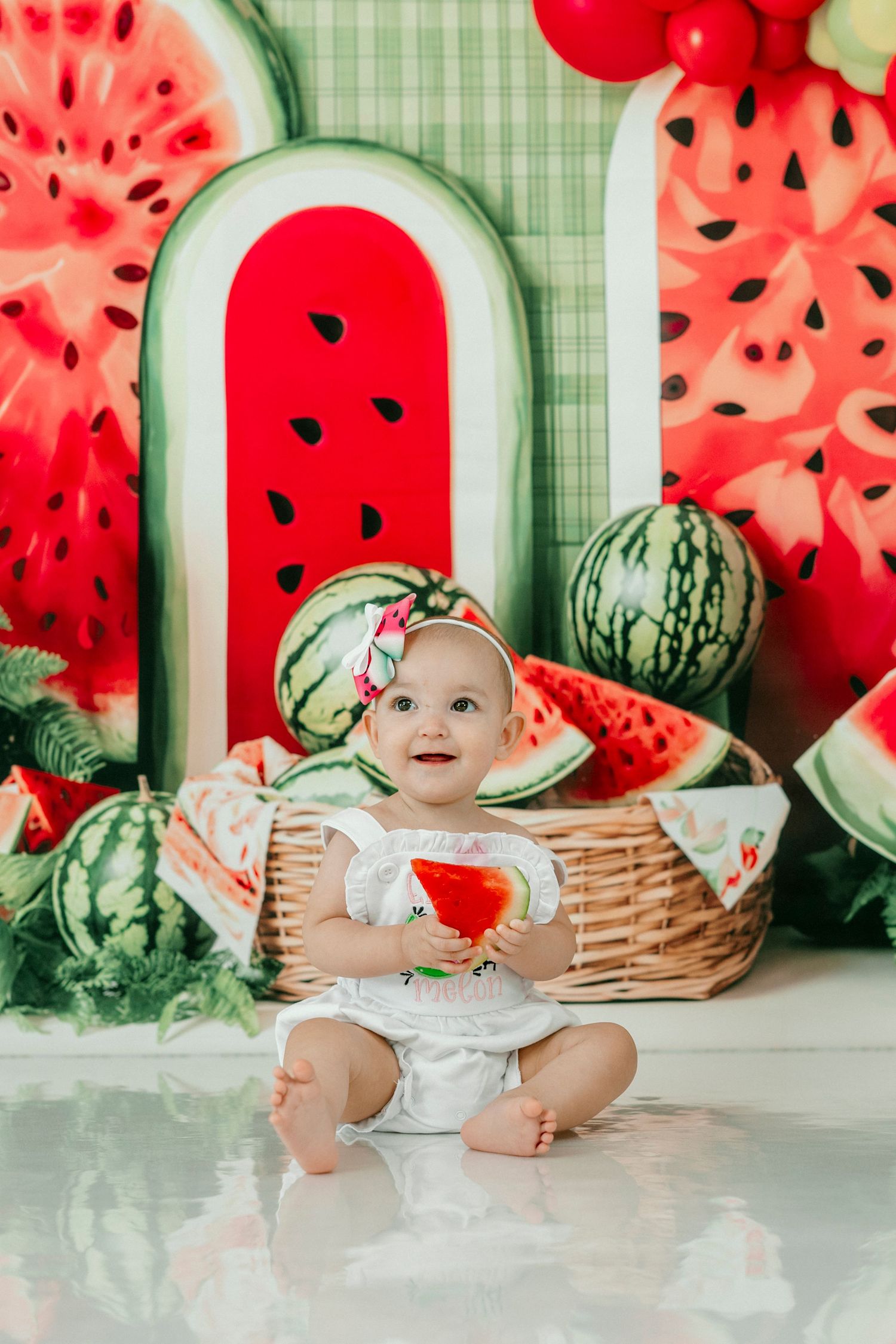 Baby girl in watermelon-themed birthday setup with decorations, balloons, and a smash cake plate.