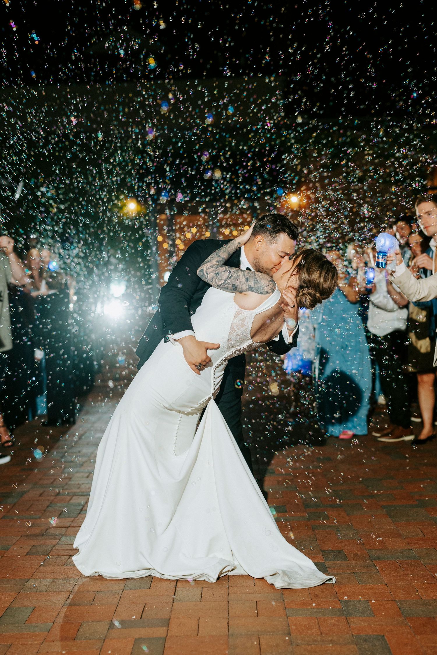 A couple shares a romantic dip kiss surrounded by colorful confetti during their wedding reception exit at night.
