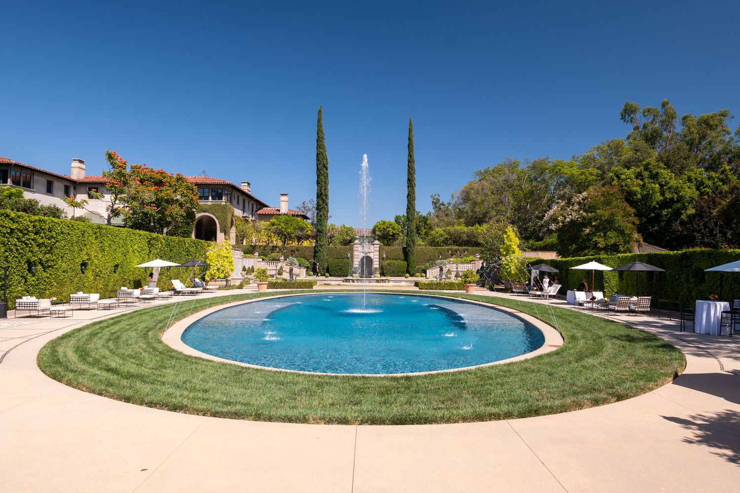 Luxury estate oval pool with fountain, lush green lawn, tall cypress trees, and elegant garden architecture under clear blue sky.