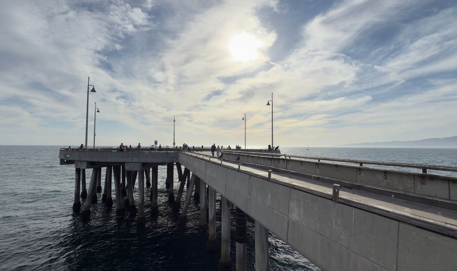 Scenic ocean pier extending over calm blue waters under a partly cloudy sky with sunlight breaking through.
