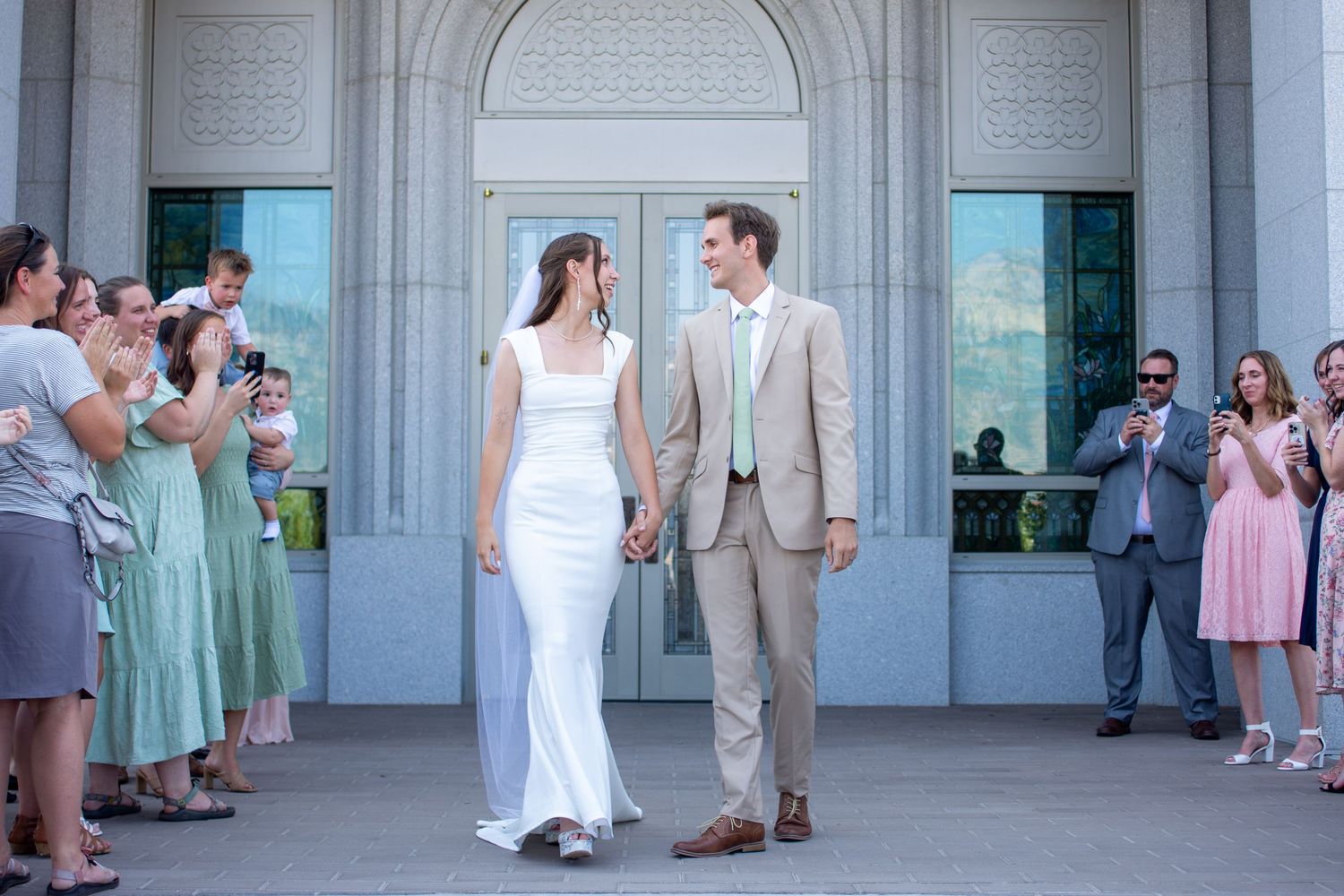 Newlywed couple holding hands outside a white wedding venue, surrounded by cheering guests in pastel attire.