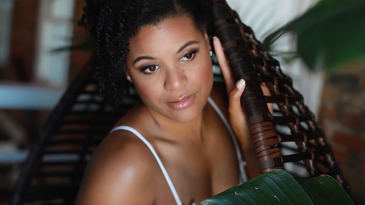 Young woman with long curly braids poses gracefully beside a tropical leaf in a serene outdoor setting.