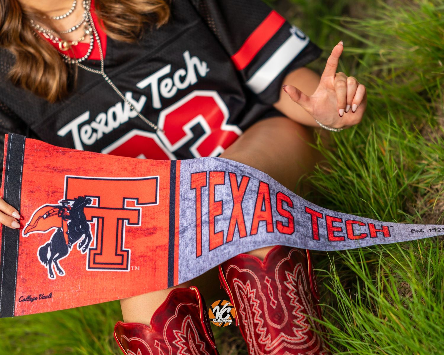 Texas Tech fan holding a red pennant flag while wearing a jersey and cowboy boots on green grass.