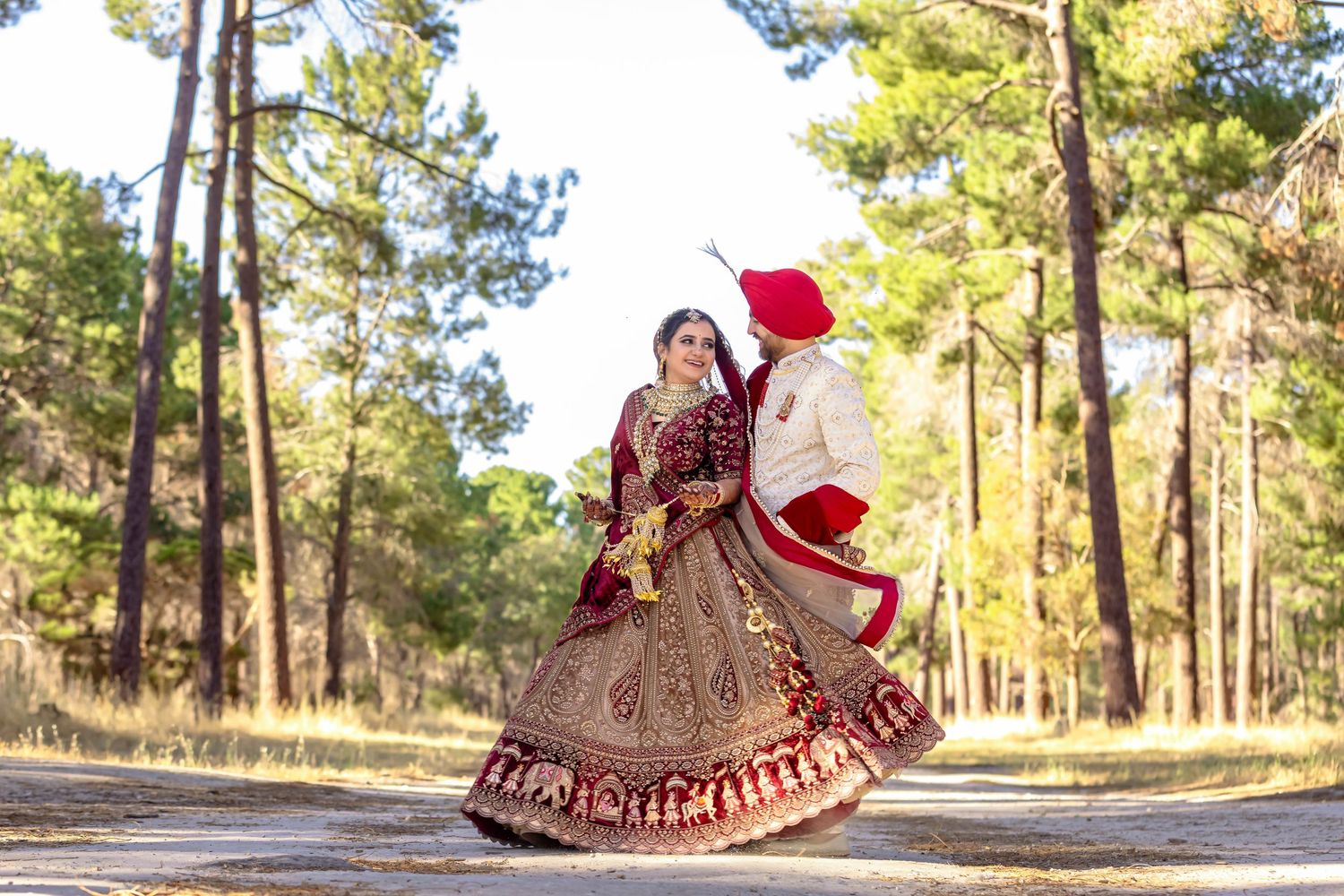 Indian bride in red lehenga twirls with groom in white sherwani among tall trees in a forest wedding photo.