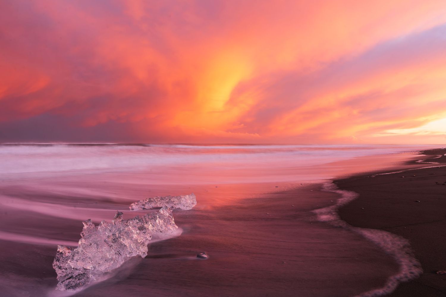 Dramatic pink and orange sunset sky over black sand beach with glacial ice chunk in Iceland.