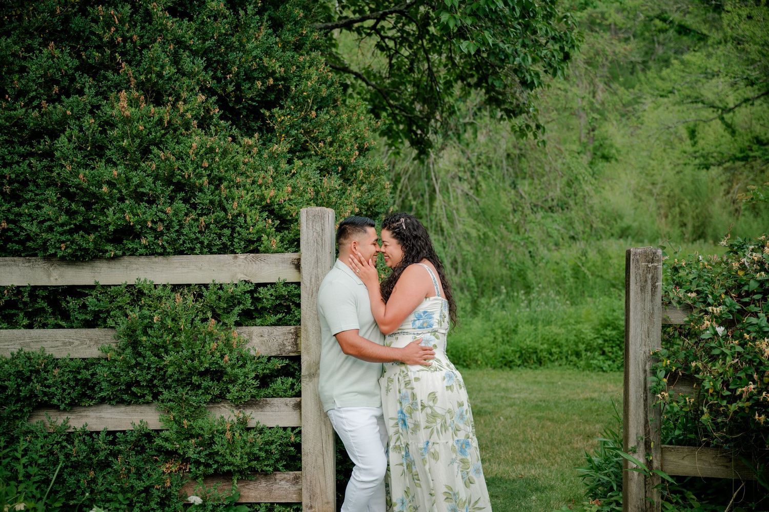 Couple sharing a romantic kiss near a rustic wooden fence surrounded by lush green trees and grass outdoors.