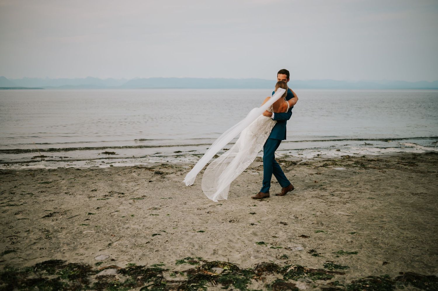 Newlywed couple on sandy beach at sunset, groom carrying bride during golden hour wedding portraits with ocean and mountains in background