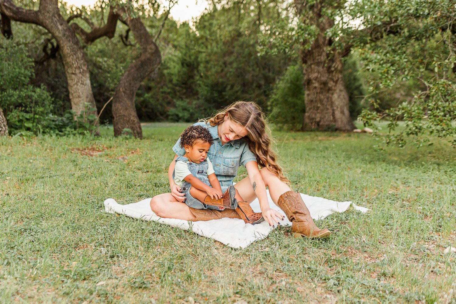Two people sit together on a white blanket in a grassy field surrounded by old olive trees.